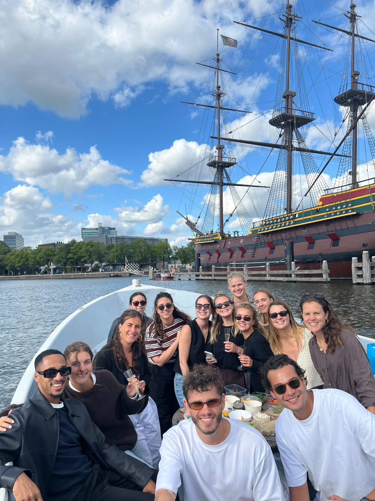 Het team van IWB op een boot in Amsterdam, met een oud zeilschip op de achtergrond